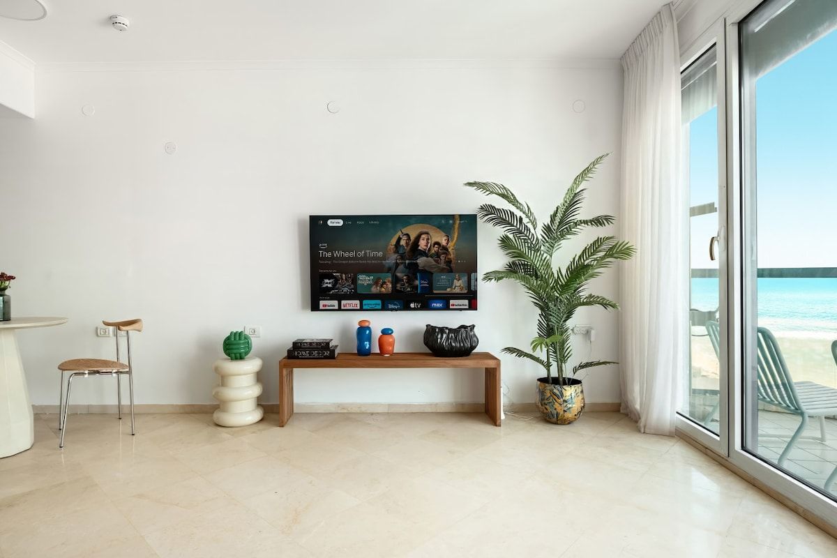 A modern living room with a flat-screen TV mounted on the wall, a wooden bench, a potted plant, and a view of the beach through large windows.