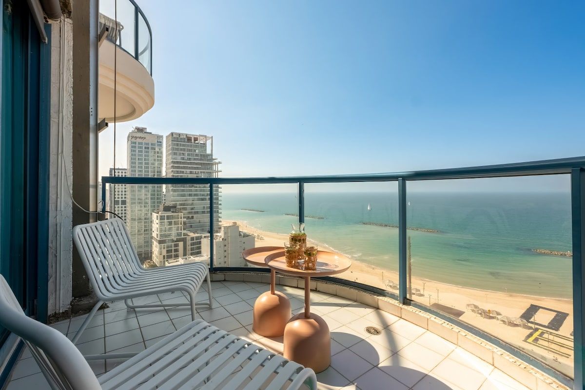 A balcony with a view of the beach, a table with drinks, and two lounge chairs.