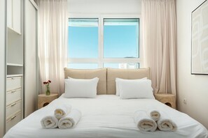 A hotel room with a bed, two bedside tables, a nightstand, a window with curtains, and a wall-mounted artwork.