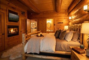 Un dormitorio rústico de cabaña de troncos con chimenea, una cama con almohadas, una mesita de noche con un libro y un televisor montado en la pared.