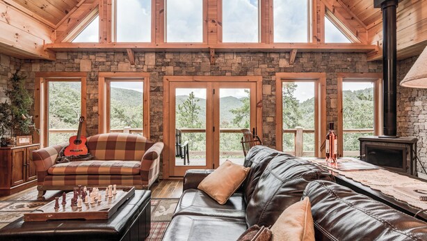 A spacious living room with a stone fireplace, a wooden ceiling, and large windows offering a view of the outdoors. - Big Sky Lodge 5 Br + Bonus-4 Ensuites, Mt Views, hot tub, game room,Pet friendly (McDowell County)