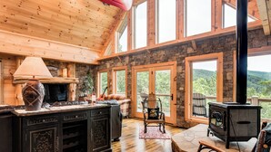 A cozy cabin interior with a stone fireplace, wooden ceiling, and large windows offering a view of the outdoors. - Big Sky Lodge 5 Br + Bonus-4 Ensuites, Mt Views, hot tub, game room,Pet friendly (McDowell County)