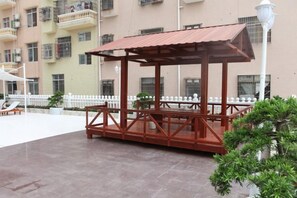 A wooden gazebo with a roof, surrounded by a white fence, in an outdoor area with a building in the background. - Dong He Hotel (Dongxing)