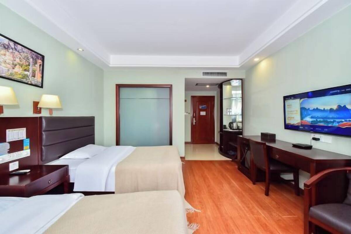 Quarto de hotel com duas camas, uma escrivaninha, uma televisão e uma porta para o banheiro.
