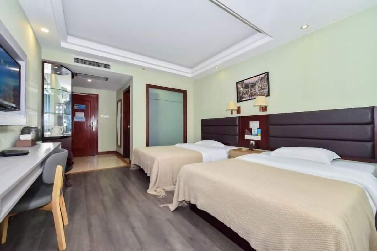 Quarto de hotel com duas camas, uma escrivaninha, uma cadeira, uma televisão e uma porta que dá para outro cômodo.