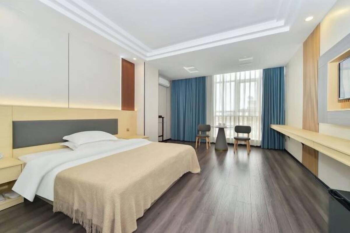 Quarto de hotel com cama, escrivaninha, duas cadeiras, janela com cortinas e iluminação embutida no teto.