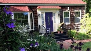 A house with a red roof, a purple door, and a front porch with steps. There are potted plants and flowers in the front yard.