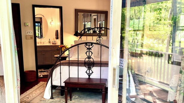A bedroom with a bed, a nightstand, a mirror, and a window with a view of greenery.