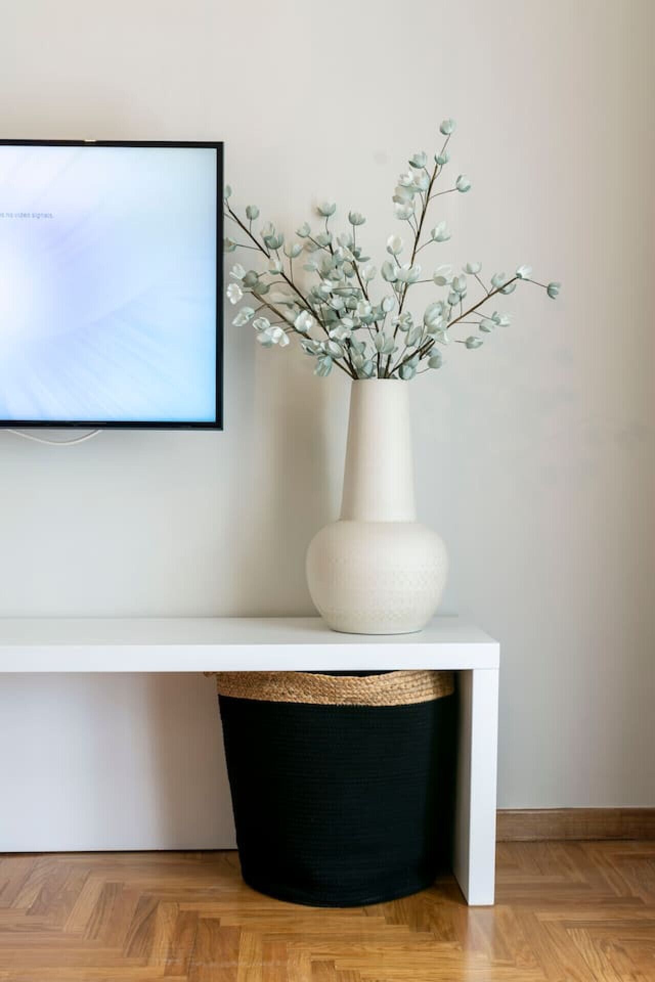Ein weißer Konsolentisch mit einer weißen Vase, die einen Zweig mit grünen Blättern enthält, einer schwarzen Mülltonne und einem gerahmten Bild an der Wand.