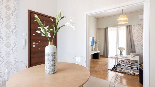 A modern hotel room with a round wooden dining table, clear plastic chairs, a white vase with white flowers, and a wooden door.