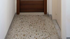A hallway with a wooden door featuring a small window and a doorbell. The floor is speckled, and there is a light fixture above the door. - Kosmos Serenity - 1BR, Central Athens (Athina)