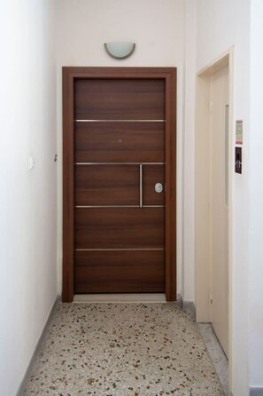 A hallway with a wooden door featuring a small window and a doorbell. The floor is speckled, and there is a light fixture above the door.