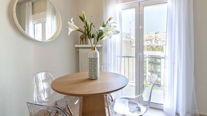 A modern room with a round wooden table, clear plastic chairs, a vase with flowers, a round mirror, and a view of a cityscape through a balcony with white curtains.