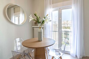 Et moderne værelse med et rundt træbord, transparente plaststole, en vase med blomster, et rundt spejl og udsigt over bybilledet fra en balkon med hvide gardiner.