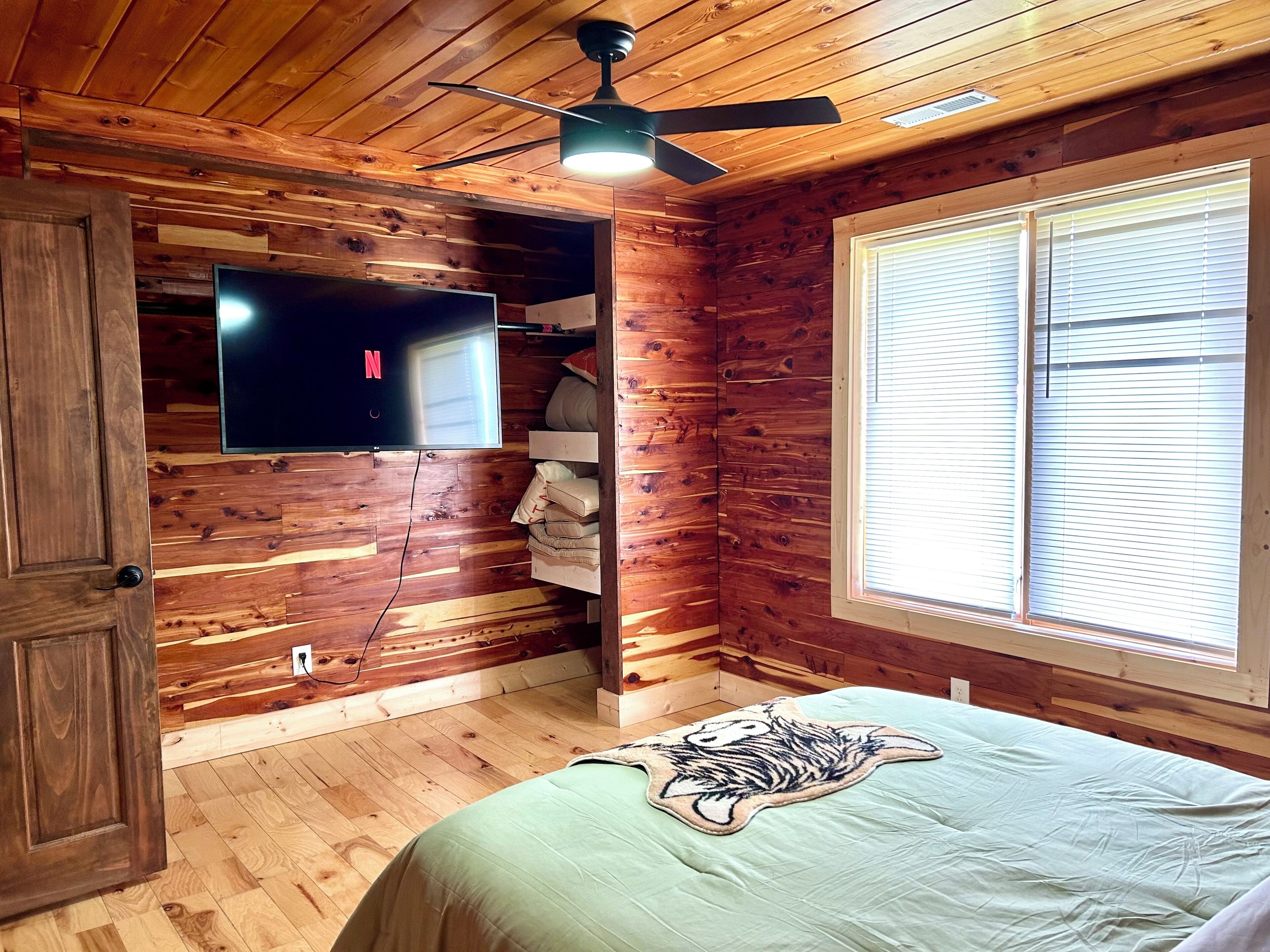 Ein Zimmer in einer Holzhütte, mit einem Bett, einem Wandfernseher, einem Deckenventilator und einem Fenster mit Jalousien.
