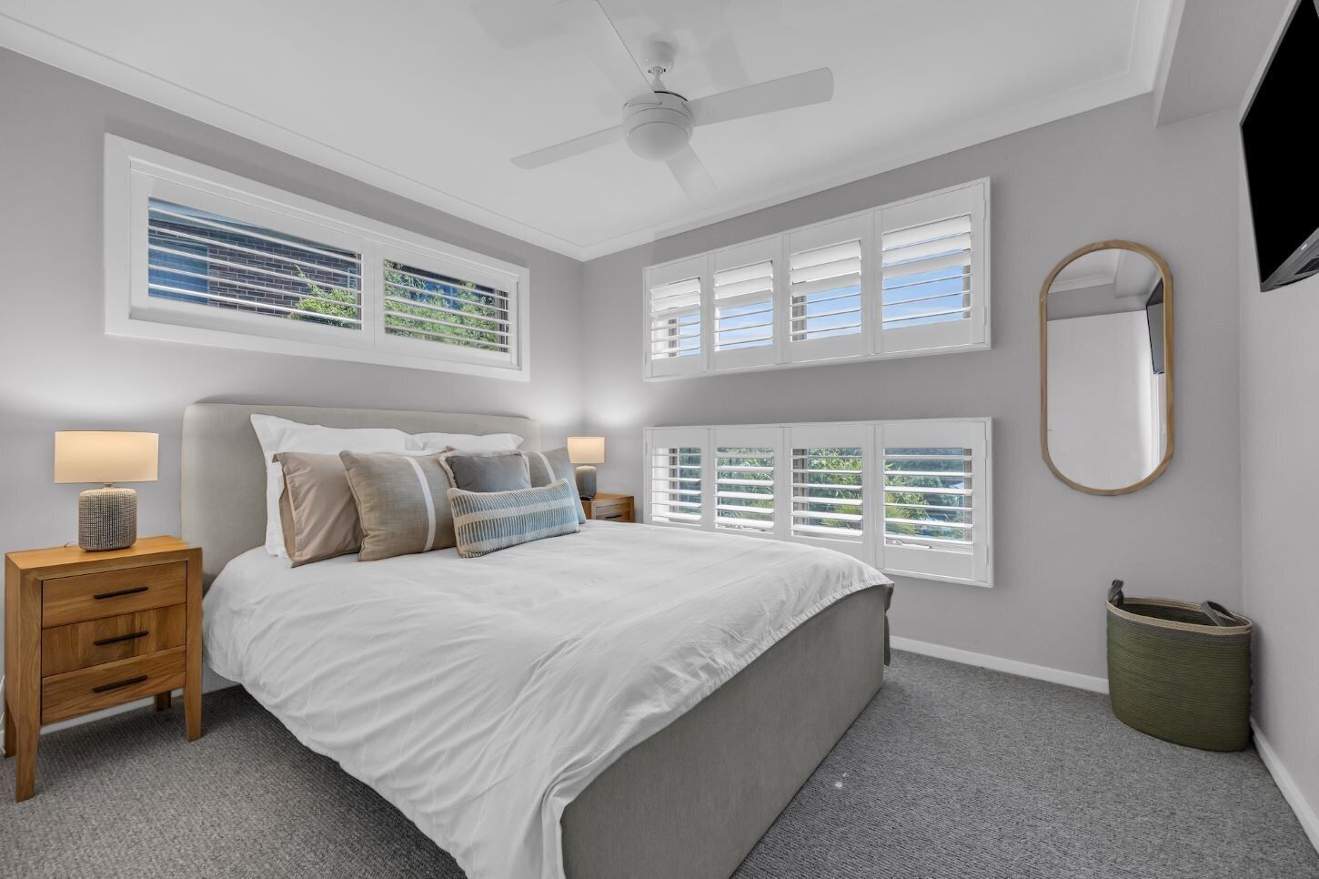 Un dormitorio con cama, mesitas de noche, ventilador de techo y ventanas amplias con persianas.