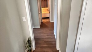 A hallway with a wooden floor, a potted plant, and a bed in the distance.