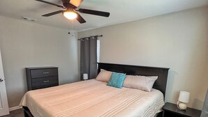 A bedroom with a bed, a nightstand, a dresser, a ceiling fan, and a window with curtains.