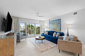 Ein modernes Wohnzimmer mit einem Flachbildfernseher, einem Deckenventilator, einem blauen Sofa, einem beigen Sessel und einem großen Fenster mit Blick auf Palmen.