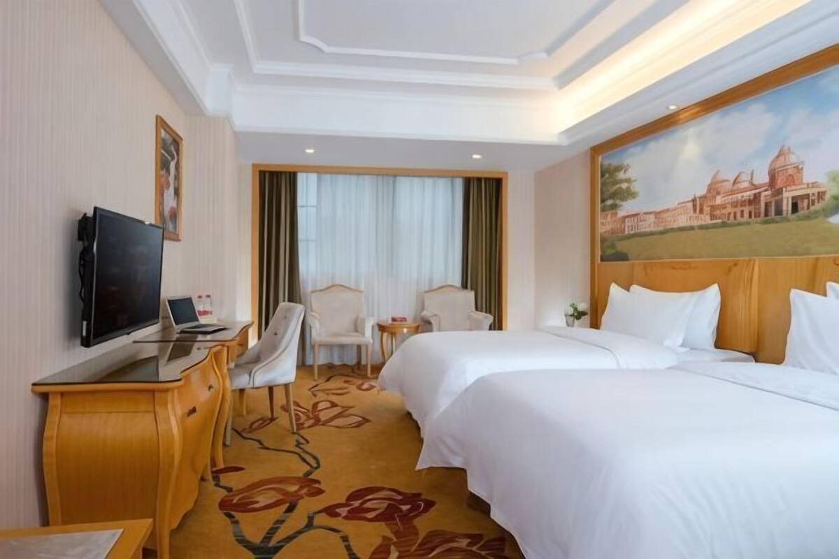 A hotel room with a bed, a desk with a TV, a chair, and a painting on the wall.