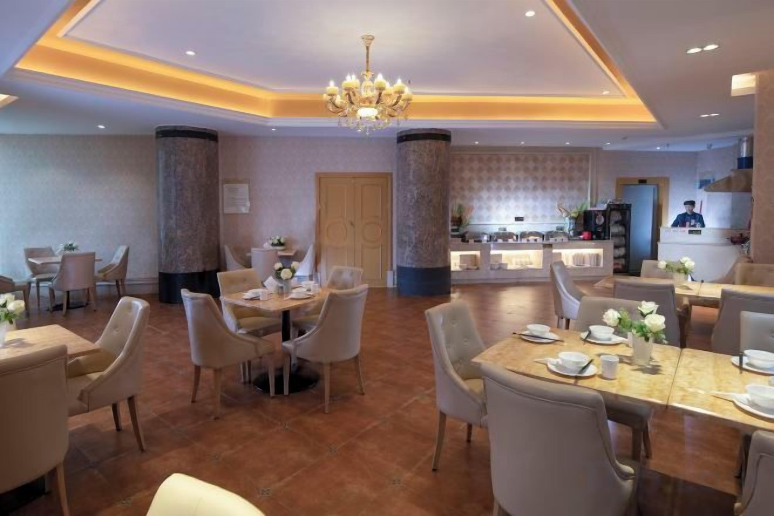 A hotel dining area with multiple tables and chairs, a chandelier, and a kitchen area in the background.