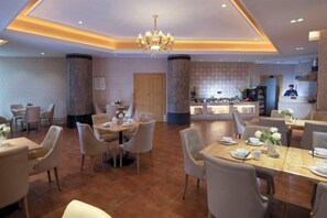 A hotel dining area with multiple tables and chairs, a chandelier, and a kitchen area in the background. - Vienna International Hotel (Chenzhou Wuling Square) (Chenzhou)