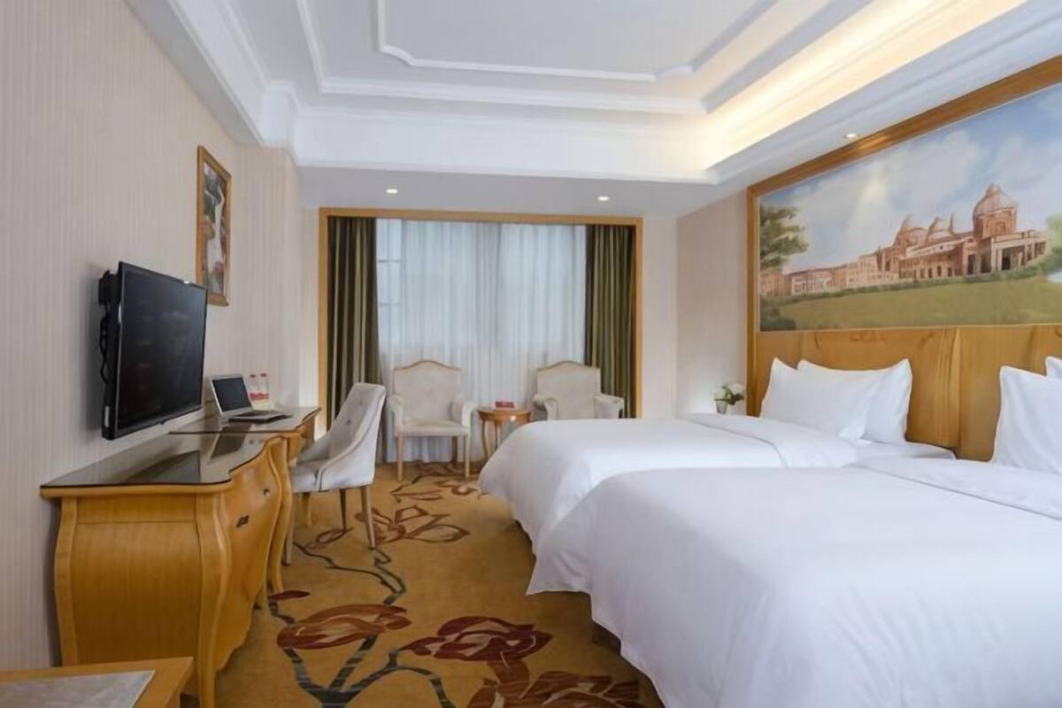 A hotel room with a bed, a desk with a computer, a TV, two chairs, and a painting on the wall.