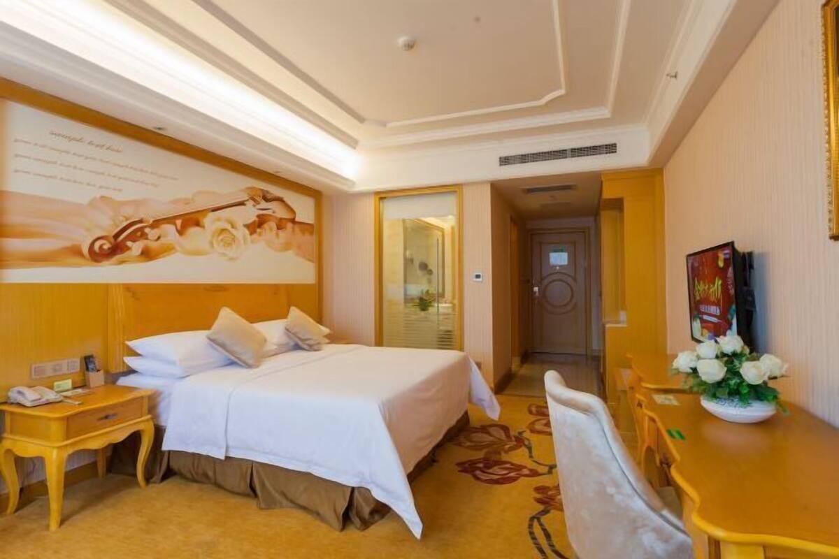 A hotel room with a large bed, a desk, and a chair. There is a painting on the wall and a window with curtains.
