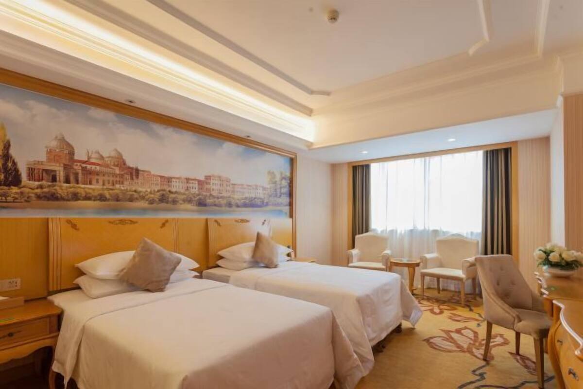 A hotel room with two beds, a painting of a cityscape, a window with curtains, and a small table with a chair.