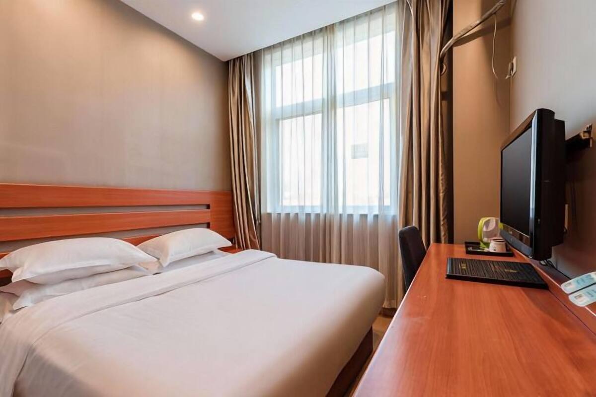A hotel room with a bed, a desk with a computer, a TV, and a window with curtains.
