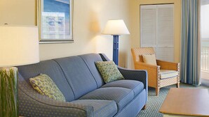Habitación de hotel con un sofá azul, una silla de mimbre, una lámpara, una mesa de madera y una ventana con cortinas.