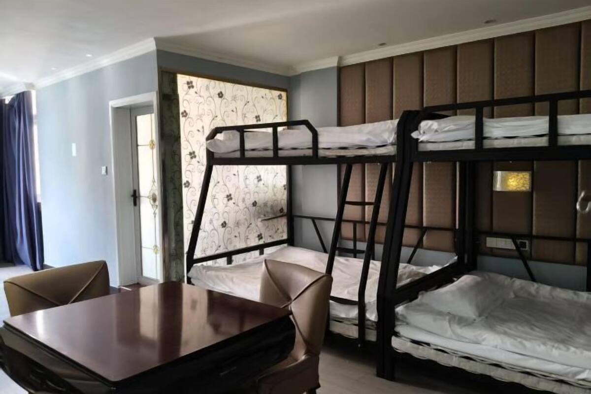 Une chambre avec un lit superposé, une table et des chaises en bois, un rideau à motifs et une fenêtre avec des stores.