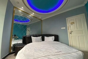 Un dormitorio con una cama grande, un espejo, una puerta blanca e iluminación azul en el techo.