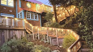 A wooden house with a staircase leading to a deck, surrounded by greenery. - Romantic Waterfront/Fire Pit/Hot Tub/Mins. to Beaches & Restaurants/Pet Friendly (Bodega Bay)