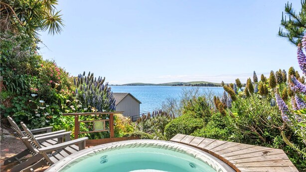 A hot tub with a wooden deck, surrounded by lush greenery and overlooking a body of water. - Romantic Waterfront/Fire Pit/Hot Tub/Mins. to Beaches & Restaurants/Pet Friendly (Bodega Bay)