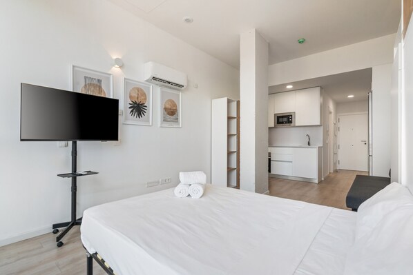 A modern hotel room with a bed, a flat-screen TV, artwork on the wall, a kitchenette, and a bathroom.