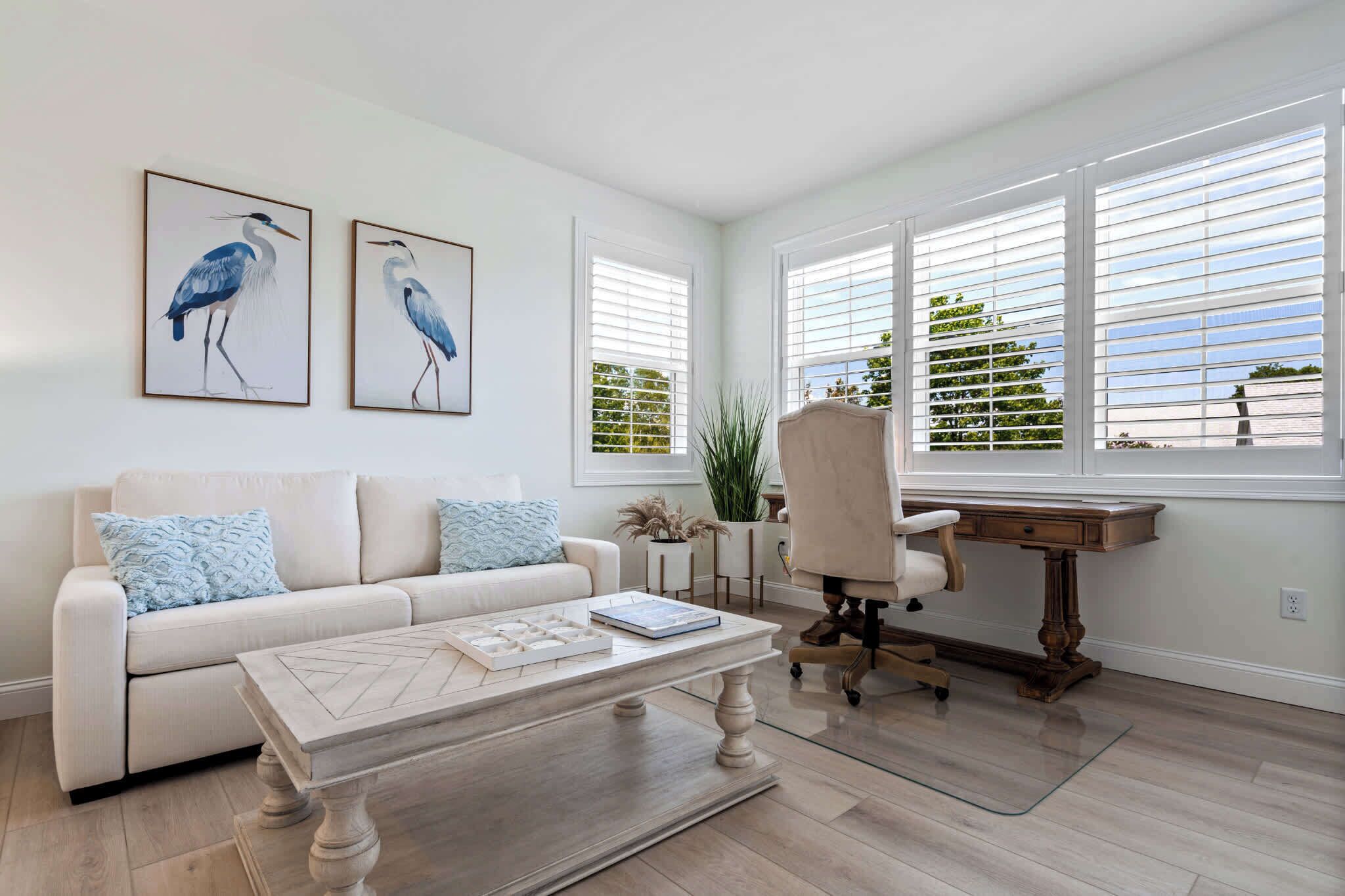 A living room with a white sofa, a wooden coffee table, a desk with a chair, and two large windows with blinds.