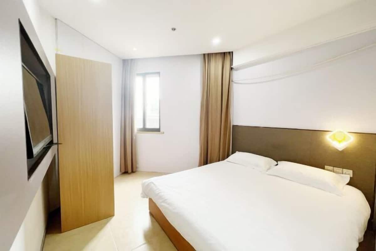A modern hotel room with a large bed, a flat-screen TV mounted on the wall, and a window with curtains.