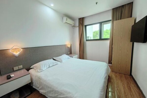 A hotel room with a bed, bedside tables, a wall-mounted TV, and an air conditioning unit.