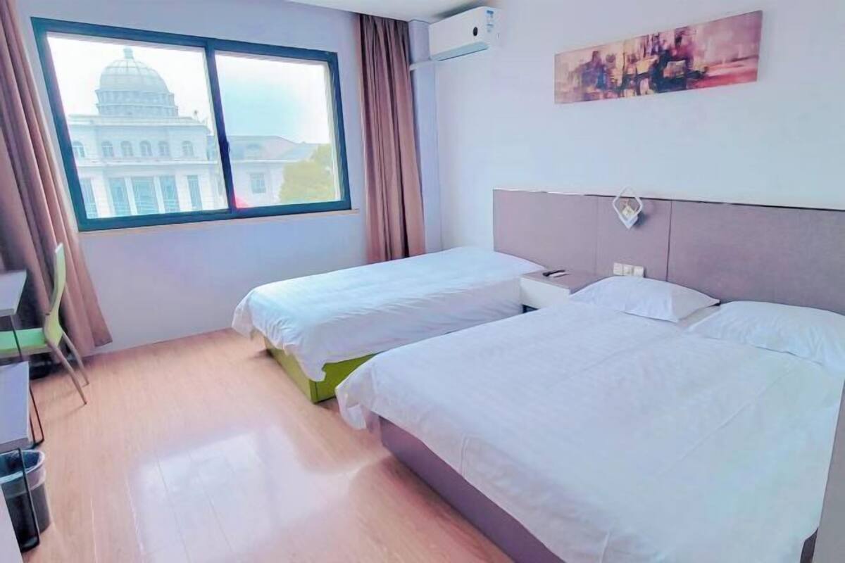 A hotel room with two beds, a desk, a chair, a window with a view of a building, and a painting on the wall.