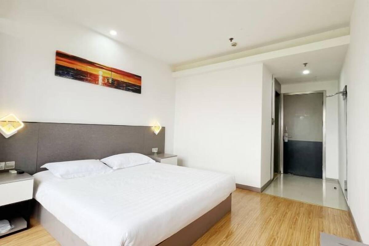 A modern hotel room with a large bed, bedside tables, a painting on the wall, and a door leading to a bathroom.