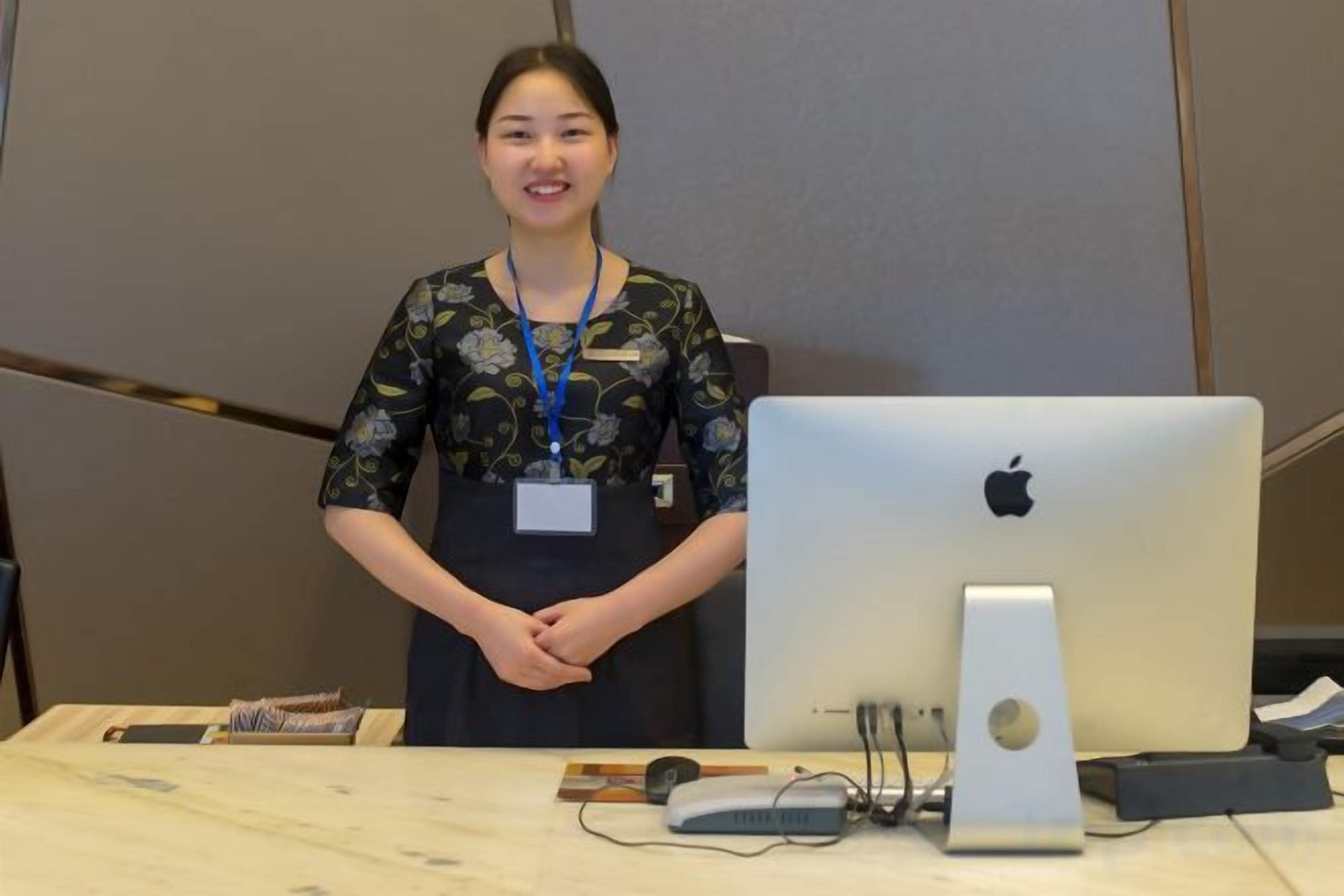 A person standing behind a desk with an Apple iMac, a microphone, and some papers.