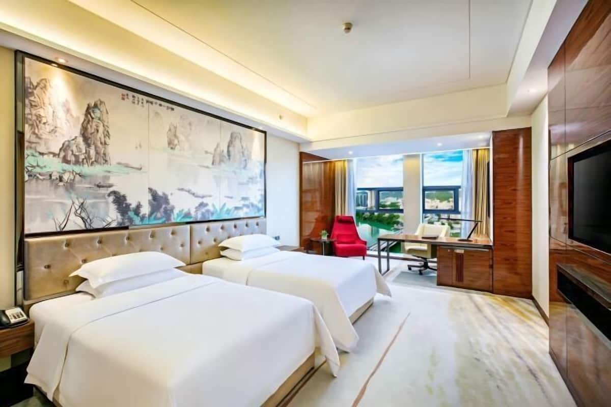 Habitación de hotel con dos camas, un escritorio, una silla, un cuadro grande, televisión y vistas al exterior.