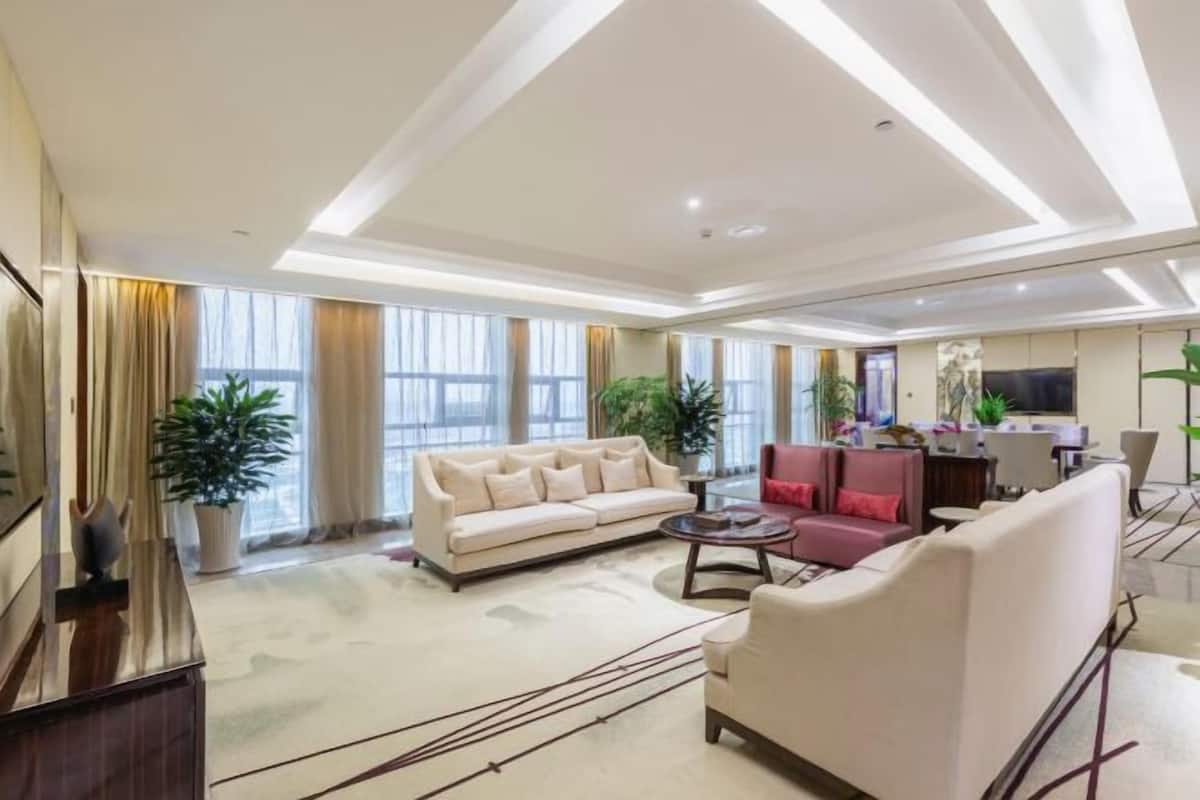 Un salón amplio con un sofá color beige, un sillón rojo y una mesa de centro. Se observan ventanas grandes con cortinas, un televisor de pantalla plana y un comedor con una mesa y sillas.