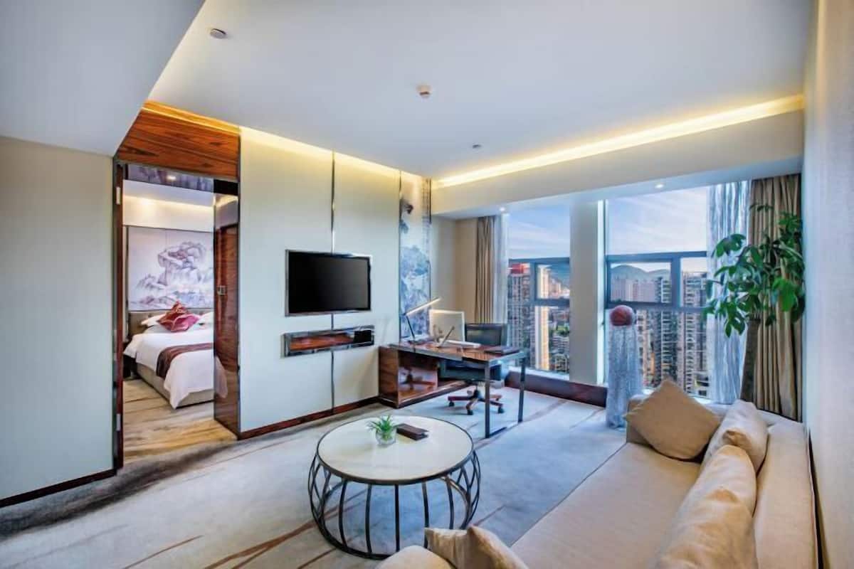 Habitación de hotel moderna con una cama grande, televisor, escritorio y sofá. Se ve un paisaje urbano a través de amplias ventanas.
