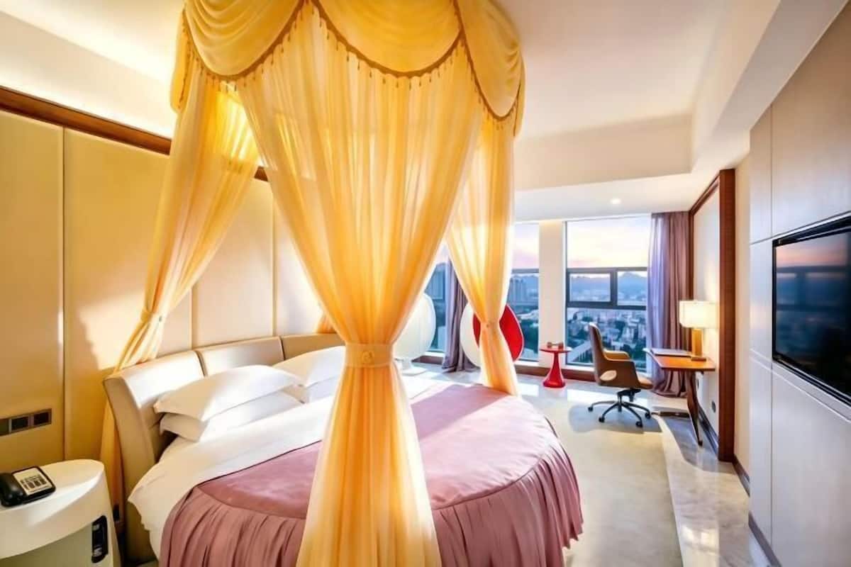 Habitación de hotel con cama, escritorio, silla, televisor y un amplio ventanal.