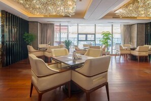 Comedor de hotel con una mesa grande y múltiples sillas, rodeado de ventanas amplias y un candelabro decorativo.