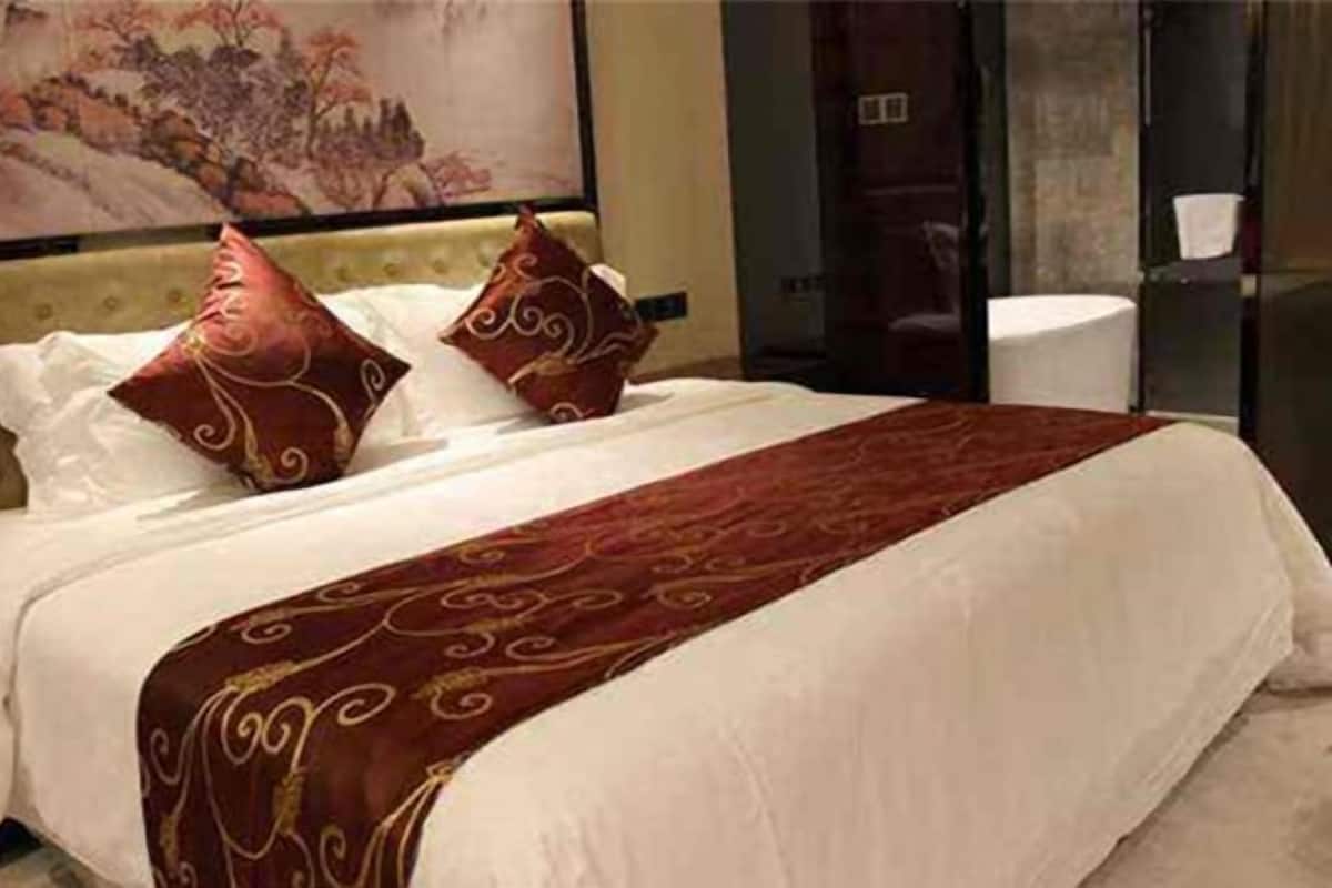 Habitación de hotel con una cama grande, dos almohadones decorativos, una colcha roja y dorada, y un cuadro en la pared.