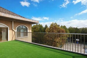 Varanda residencial com grama sintética, guarda-corpo de metal e vista para árvores e quadra de tênis.