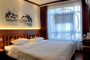 A hotel room with a bed, a wall-mounted painting, a large window with curtains, and a wooden headboard.